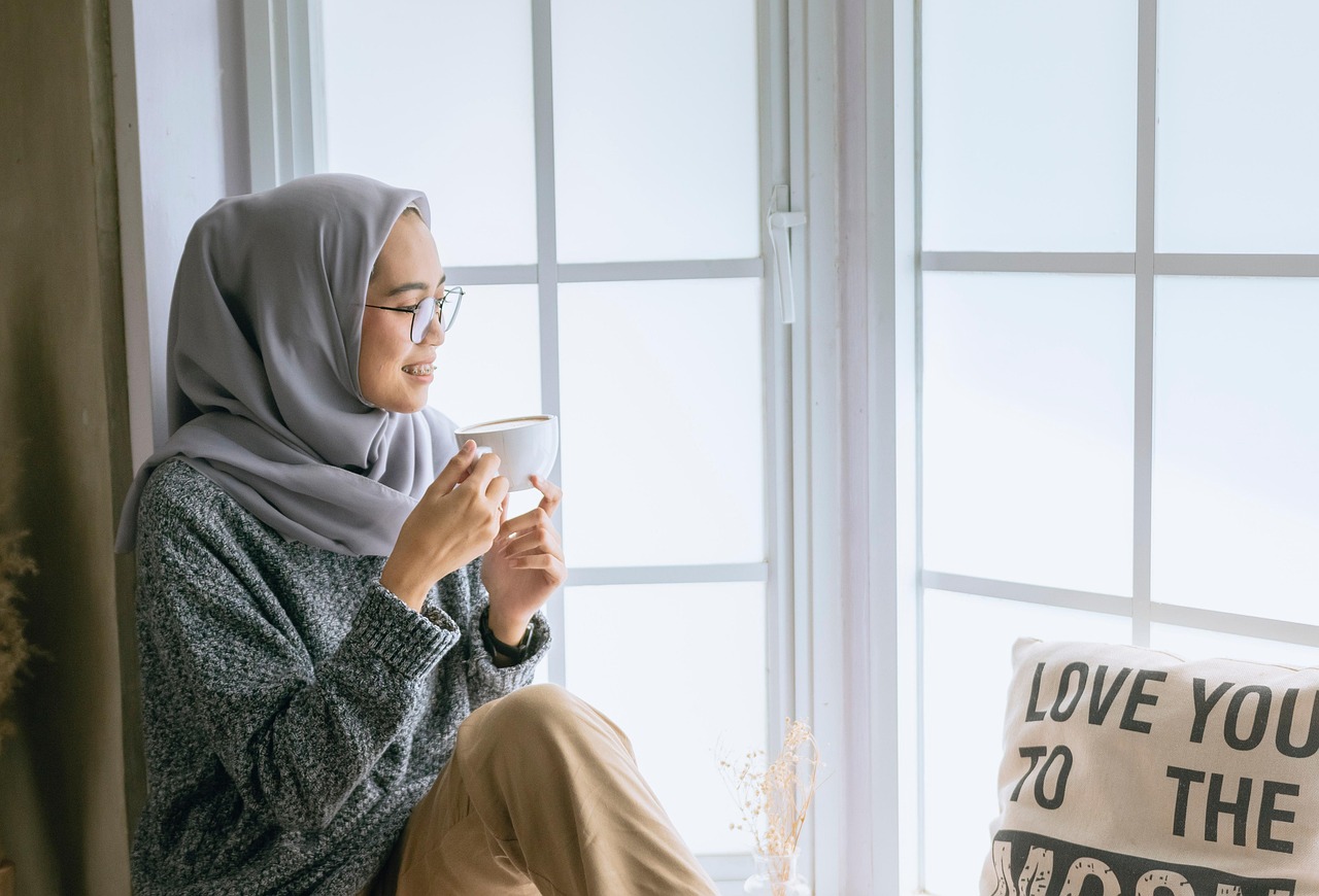 Zelfzorg bij levend verlies: een vrouw met een hoofdoek zit ontspannen met een kop koffie of thee in de vensterbank